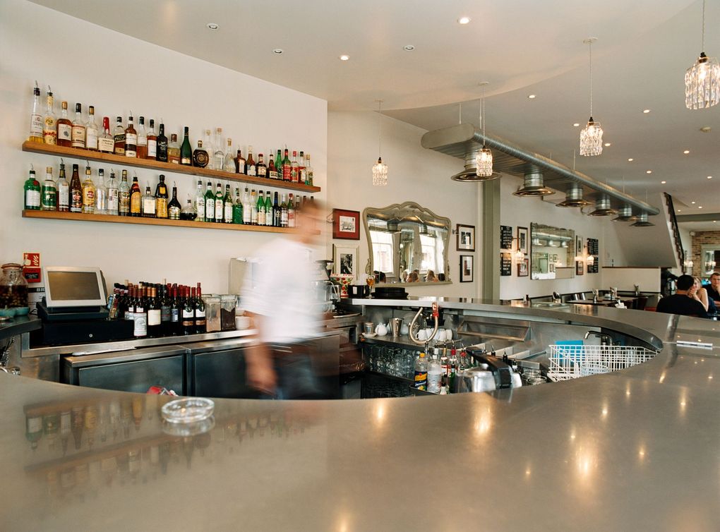 Bright modern bar interior with shelves of bottles and a blurred bartender behind the counter