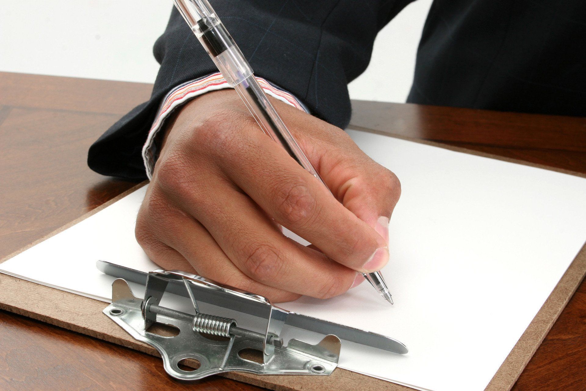 Hand writing on a clipboard, wearing a suit jacket. Pen held over white paper.