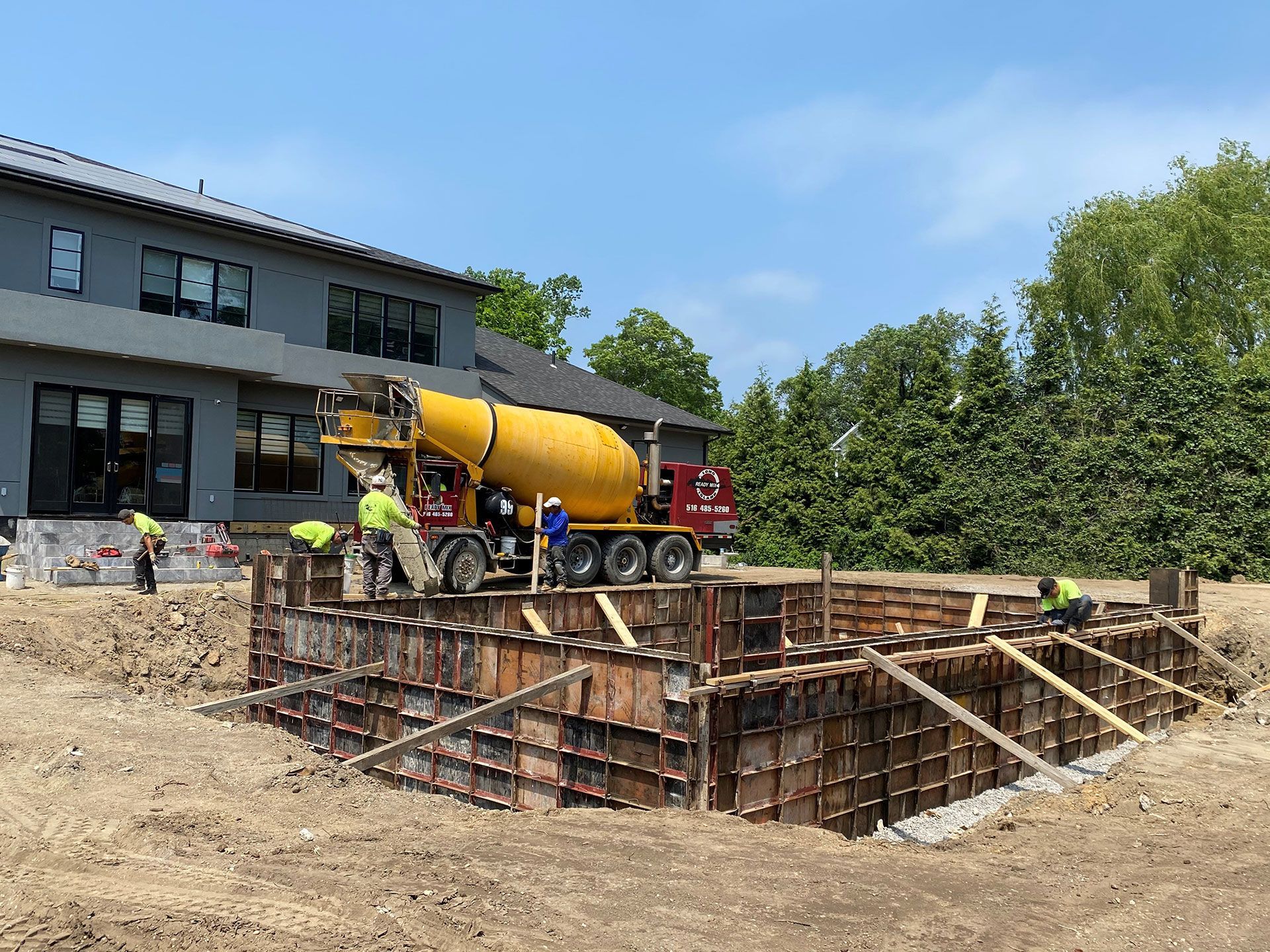 Concrete truck pouring concrete into wooden forms at a construction site near a house; workers in safety vests.