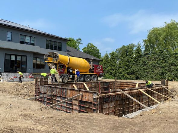 Concrete truck pouring concrete into wooden forms at a construction site near a house; workers in safety vests.