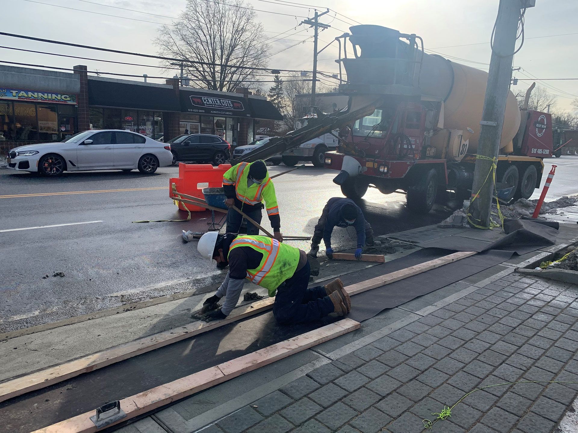 Construction workers pouring concrete at a sidewalk with a cement truck in the background. Orange traffic barriers are nearby.