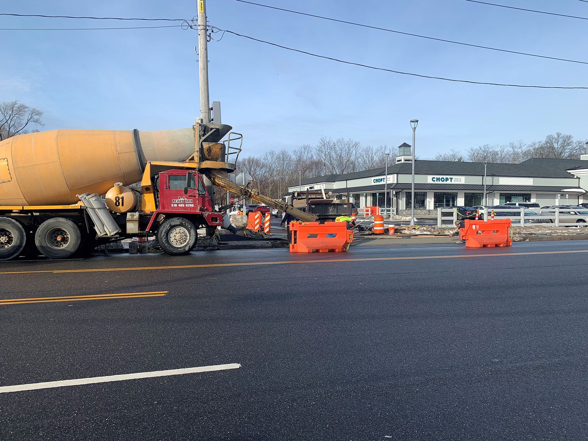 Cement truck pours concrete at a construction site near a building with workers in orange vests.