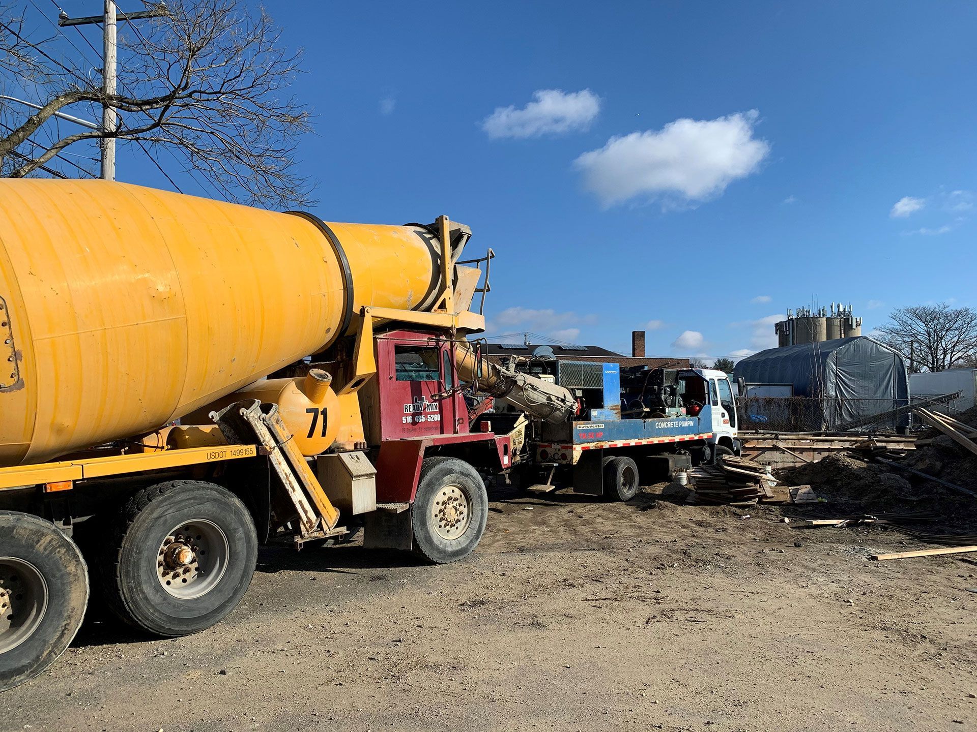 Yellow cement truck next to a smaller white truck, both on a dirt construction site under a blue sky.
