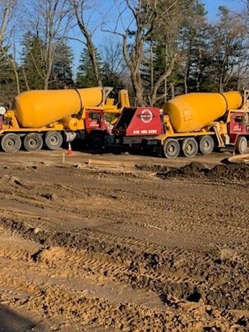 Two yellow cement trucks at a construction site, with dirt and trees in the background.