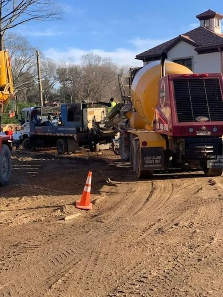 Construction site with concrete truck pouring. Brown soil, orange cone, buildings in background.