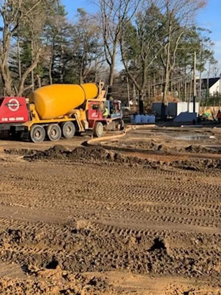 A yellow cement truck at a construction site with muddy ground and trees.