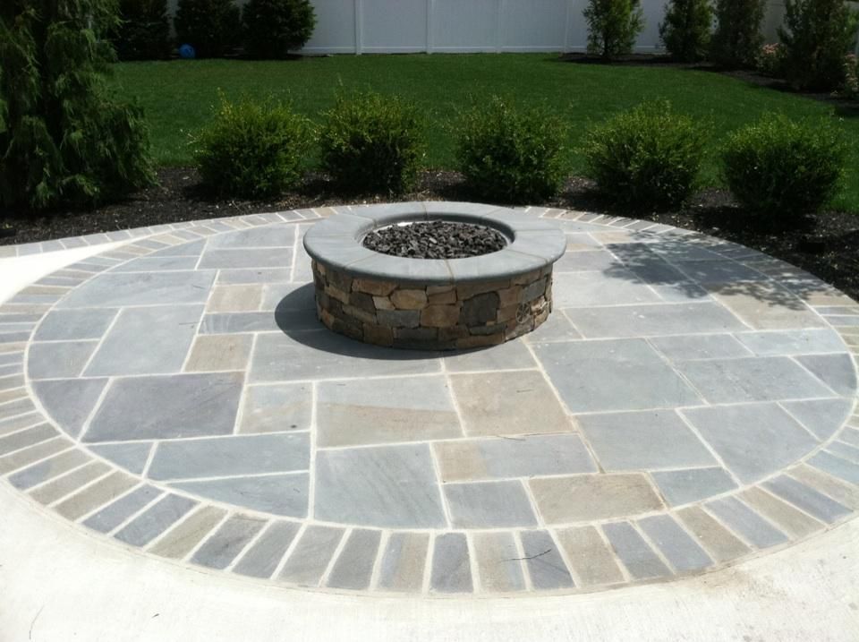 Circular stone patio with fire pit, surrounded by grass and bushes.