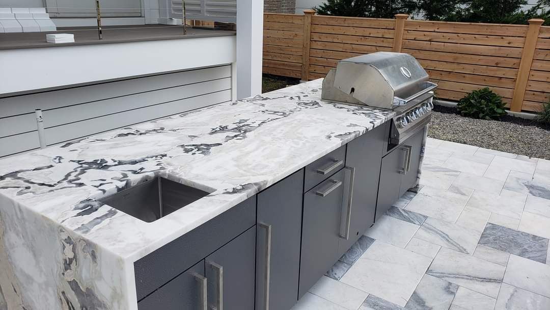 Outdoor kitchen with stainless steel grill, sink, and gray cabinetry on a marble countertop.
