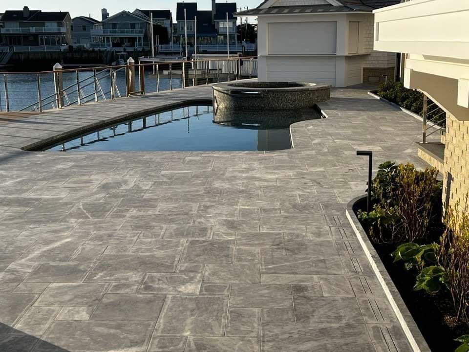 Patio with a pool and hot tub overlooking a waterway. Gray stone pavers, beige building.