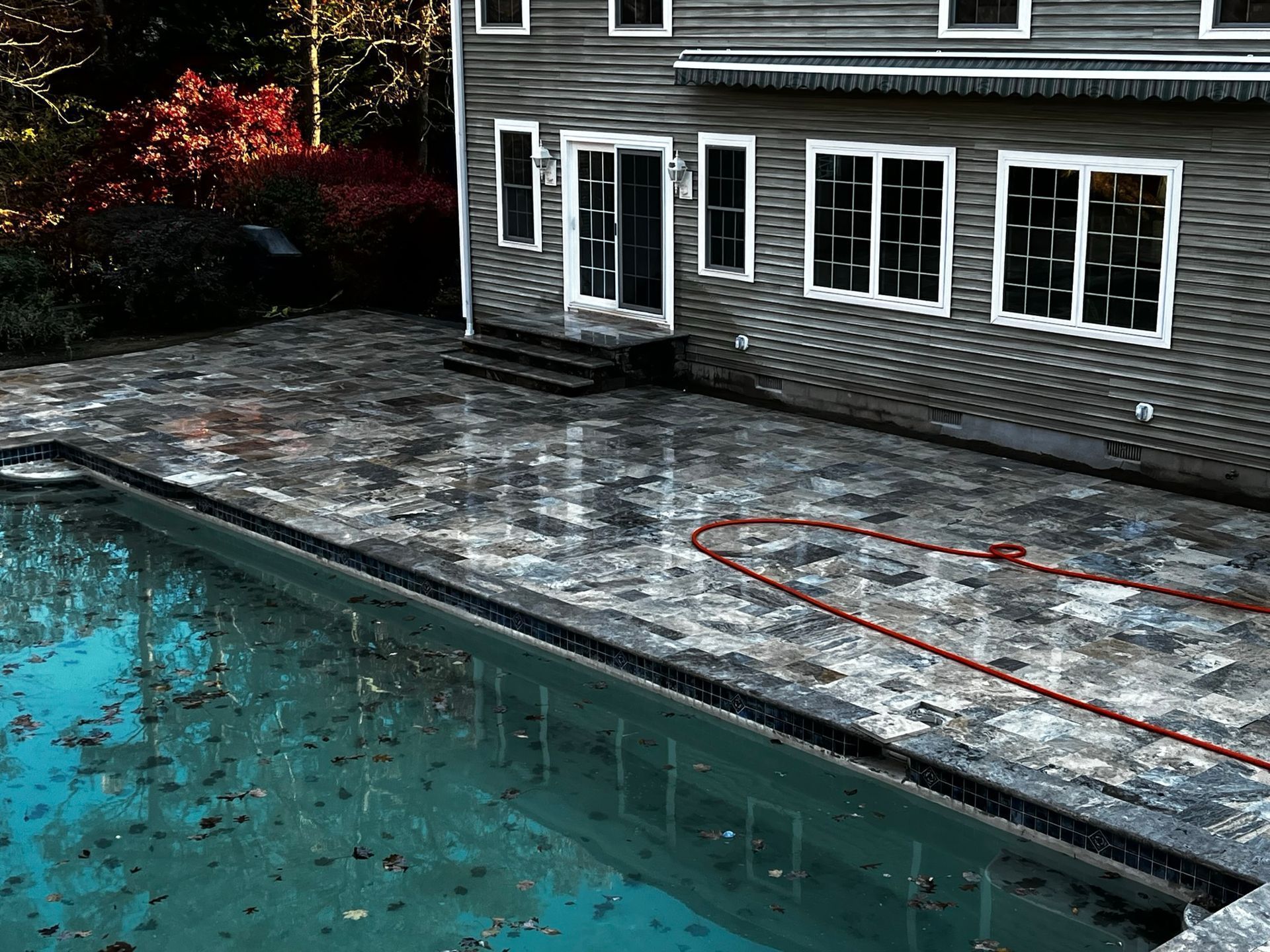 Pool with wet, stone patio and house exterior. Red hose visible.