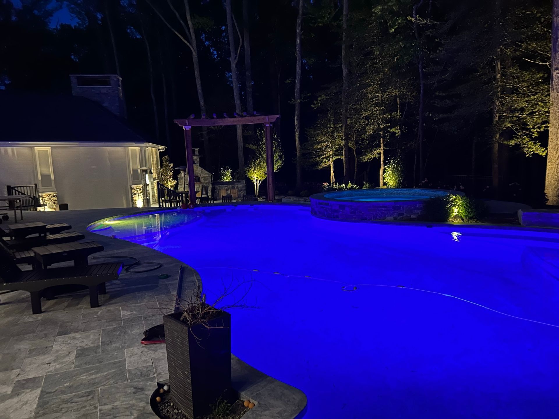 Nighttime view of a backyard pool with blue lights, illuminated by landscape lighting. A pergola and hot tub are visible.