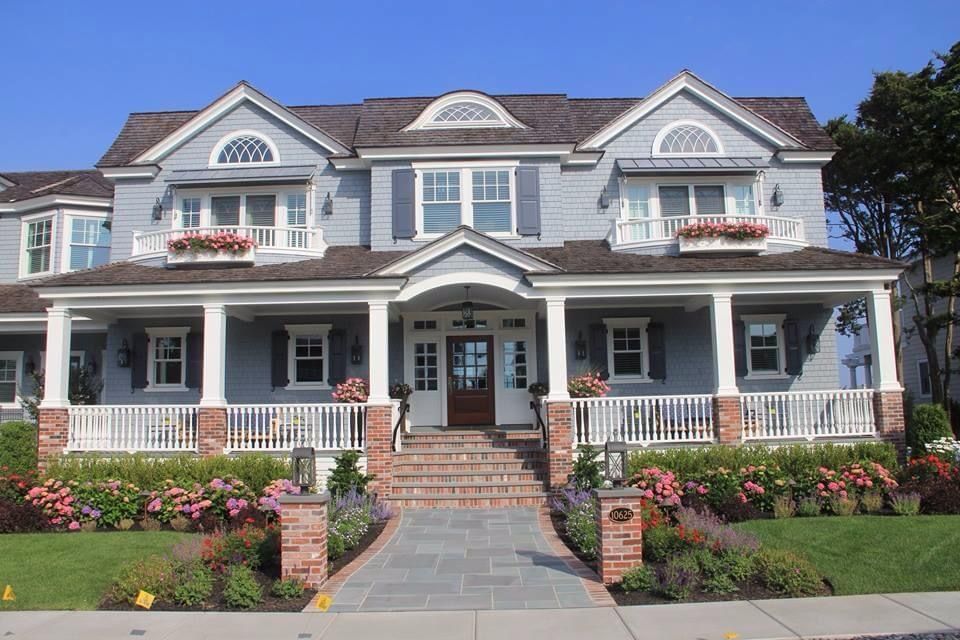 Blue-gray two-story house with porch, shutters, and landscaping, including pink flowers and a brick walkway.