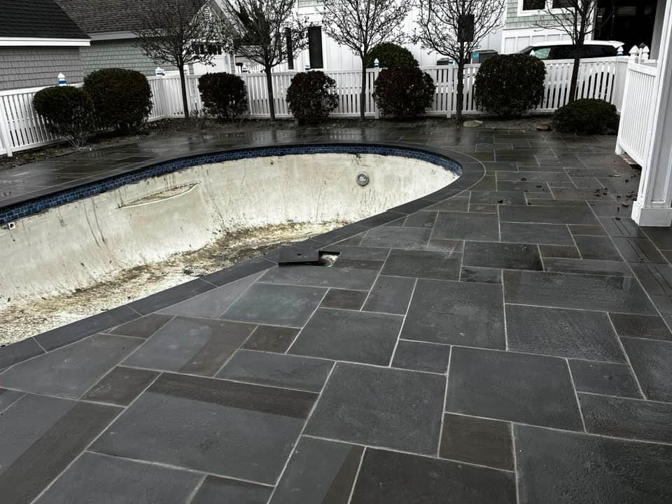 Empty pool with dark stone patio, white fence, and small bushes. Overcast day.