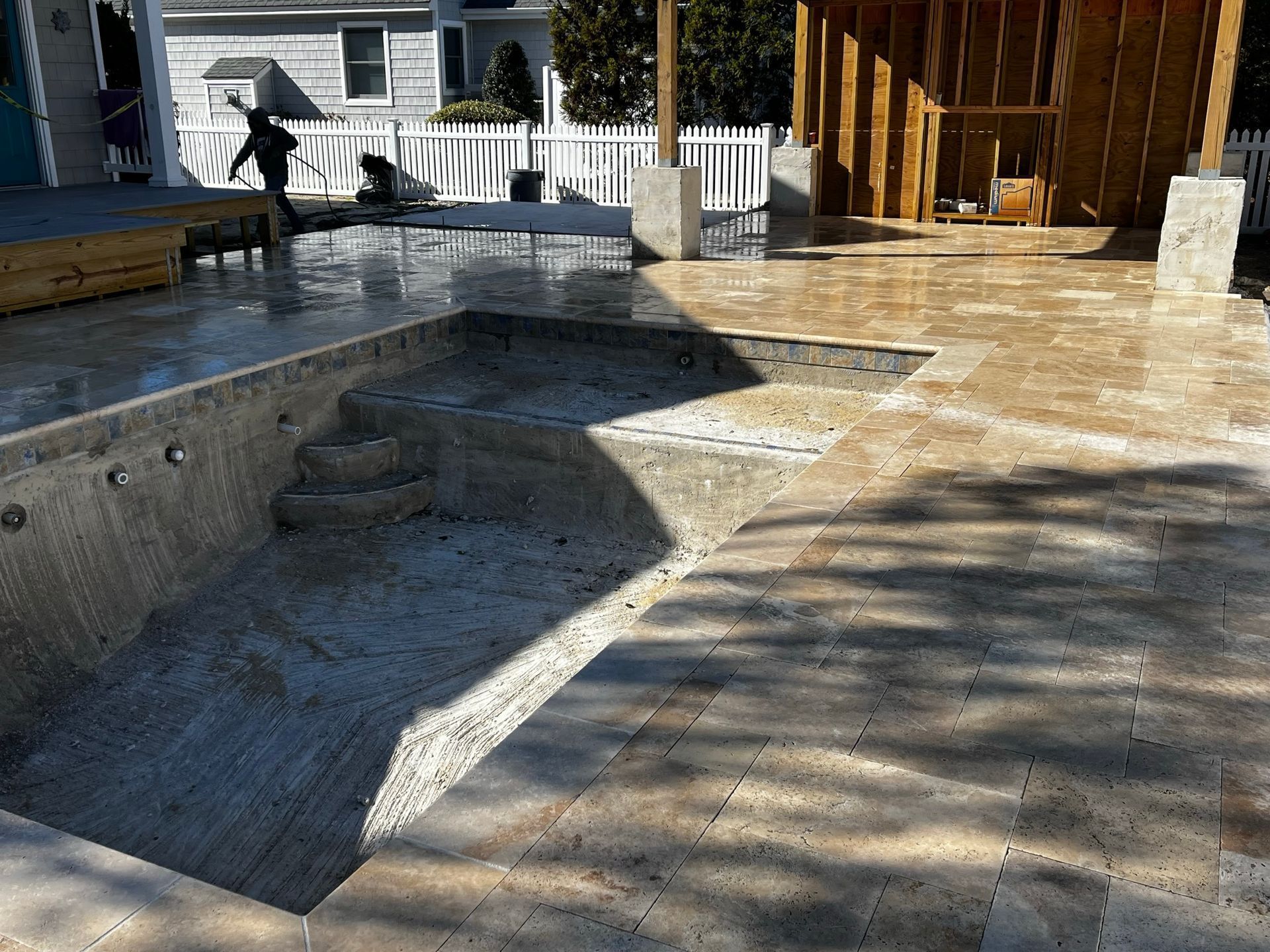 Construction of a pool and patio area. Empty pool with steps, surrounded by stone pavers. A person works in the background.