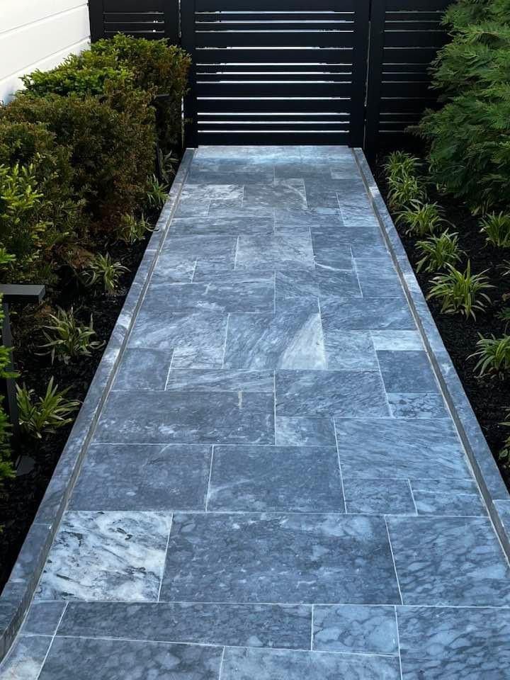 Pathway of blue-grey stone tiles leading to a black gate, flanked by green bushes and plants, black mulch border.