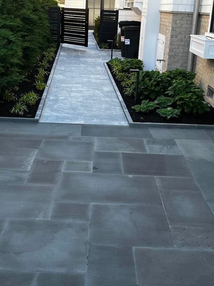 Stone patio and walkway leading to a black gate, bordered by landscaping with green plants.