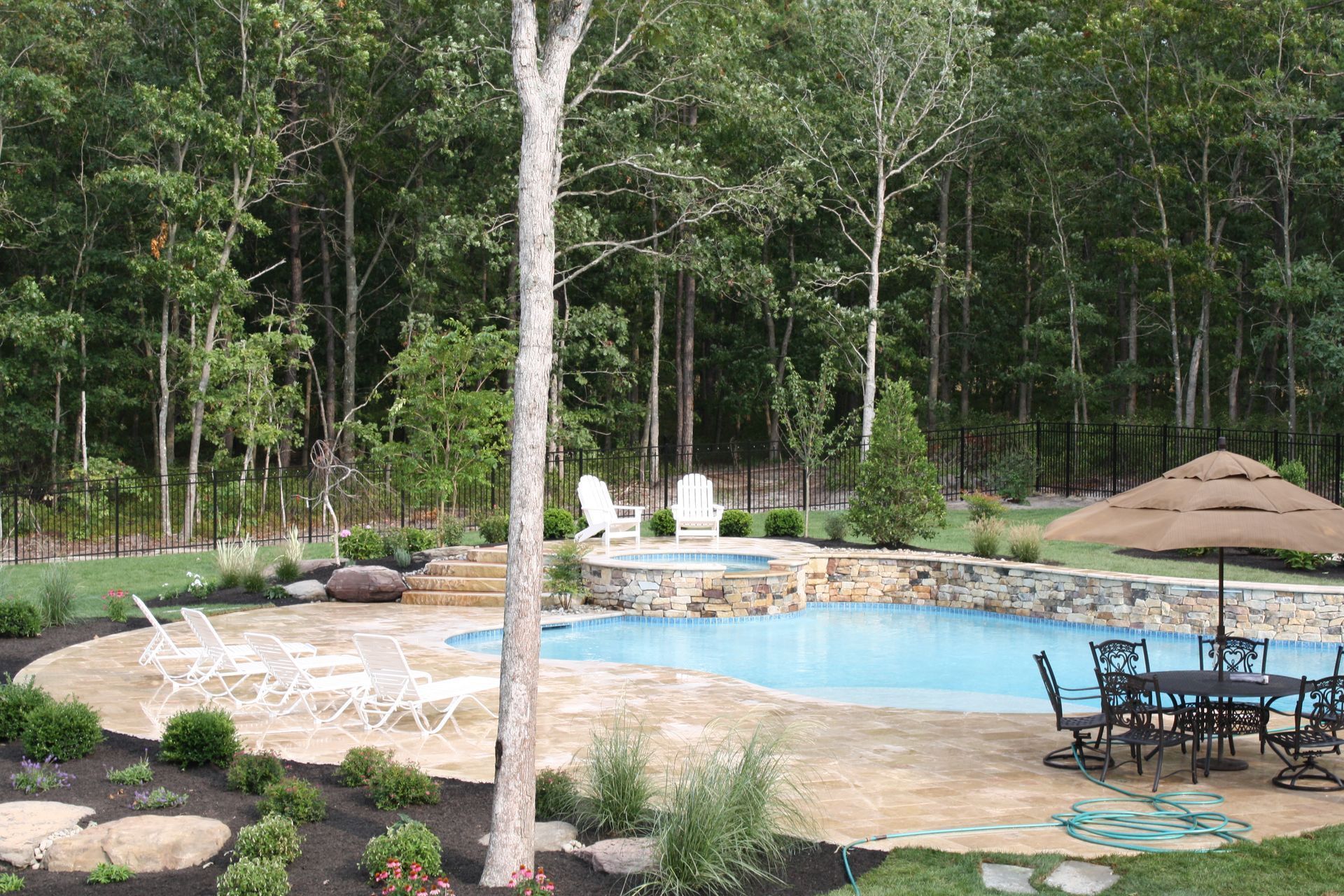 Backyard pool with stone patio, chairs, and umbrella, surrounded by trees.