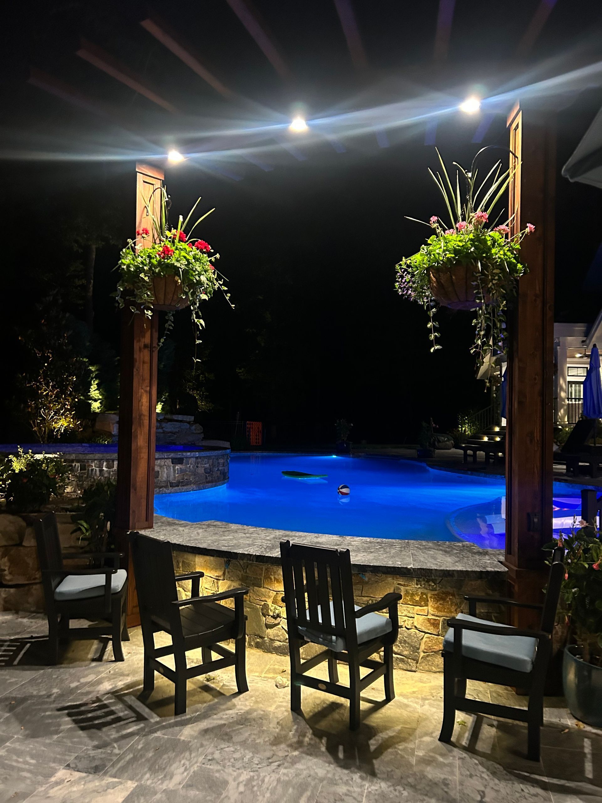 Night view of a pool with seating, hanging flower baskets, and illuminated pergola.