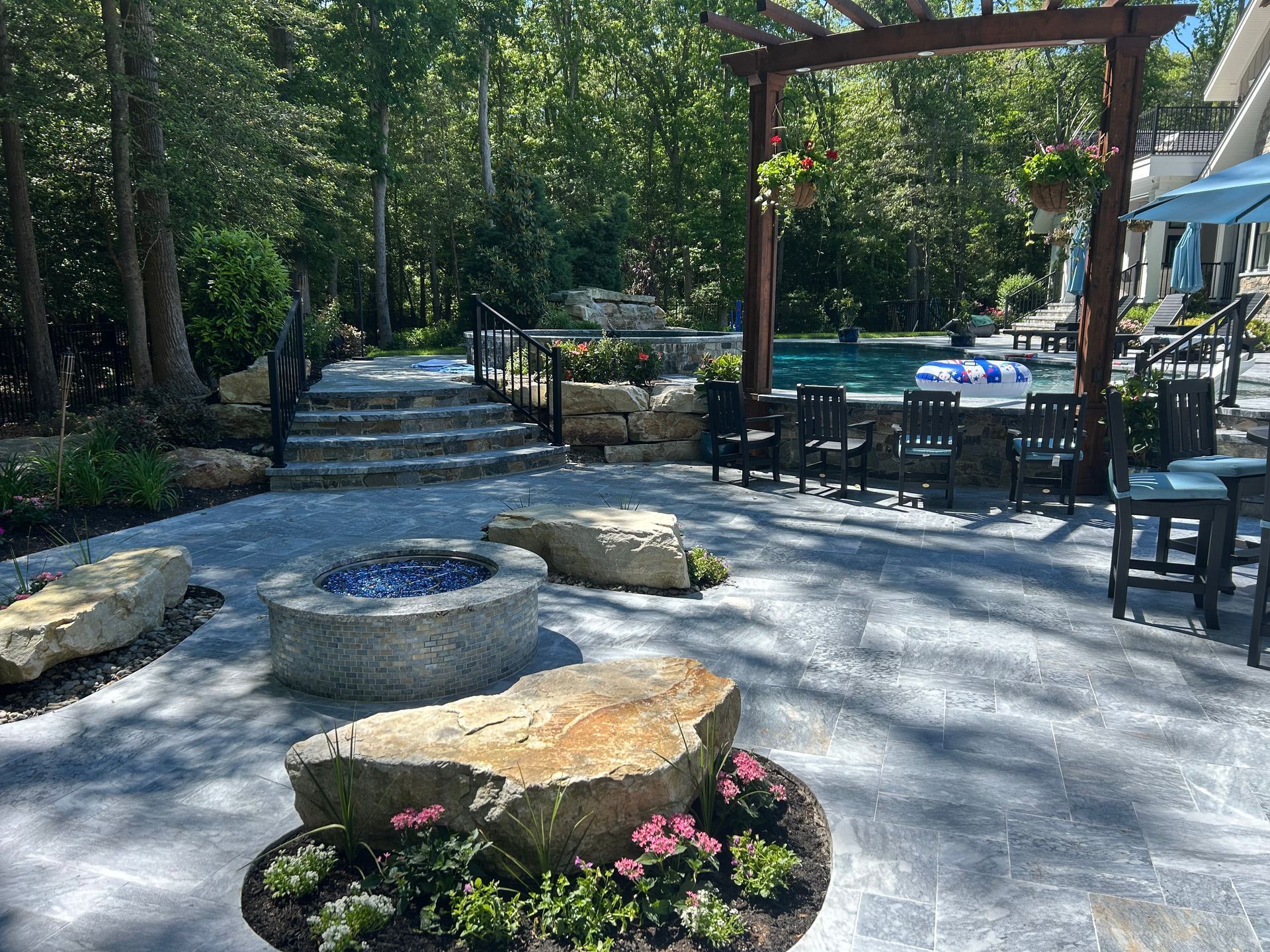 Patio with stone fire pit, seating, pool, and stairs leading to wooded area.