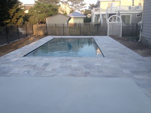 Rectangular in-ground pool surrounded by stone patio. Houses and a wooden fence are in the background.