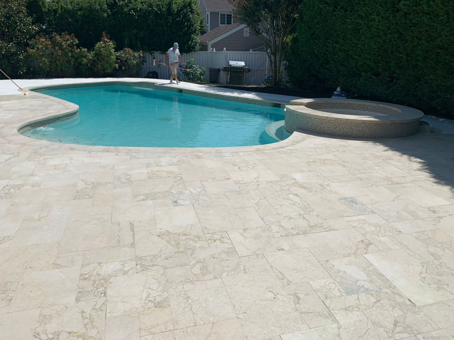 Pool with light-colored stone surround; person standing nearby. Lush green hedge in the background.