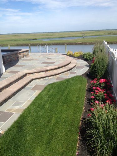 Stone patio with steps leading to waterfront. Green lawn and flower beds. Blue sky and water.