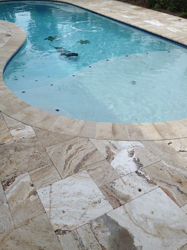 Pool with curved edge, surrounded by light-colored stone tiles. Water is clear, some debris visible. Sunny outdoor setting.