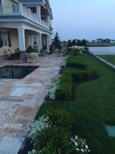 Stone patio with pool, landscaping, and house with balcony near water at dusk.