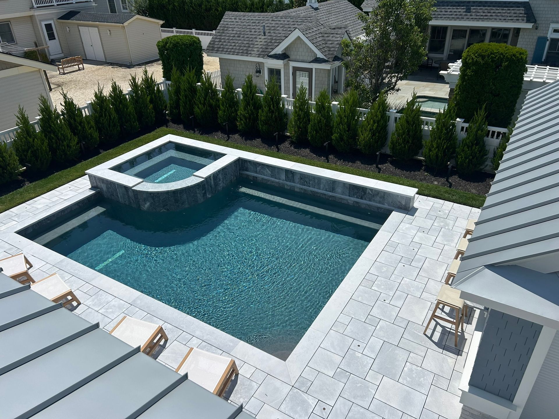 A rectangular swimming pool and attached hot tub with a paved patio. Green hedge row in background.