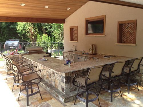 Outdoor kitchen with stone base, granite countertop, bar stools, and TV.