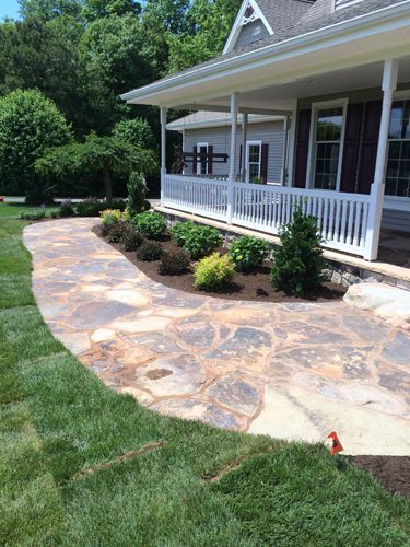 Stone pathway leading to a porch with white railing, surrounded by grass and landscaping.
