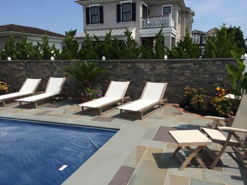 Poolside with lounge chairs, stone wall, and a multi-story house on a sunny day.