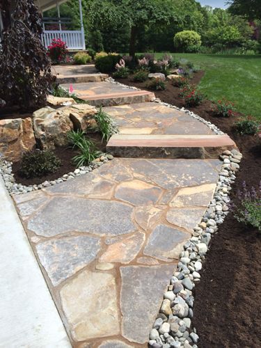Stone pathway leading up steps to a house, flanked by garden beds, rocks, and grass.