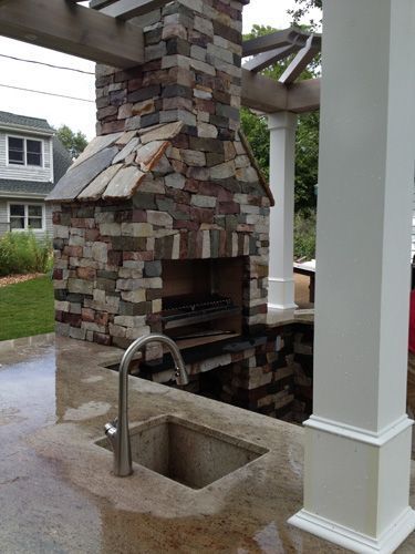 Outdoor stone fireplace with sink, under pergola; gray and tan stone, stainless steel faucet.
