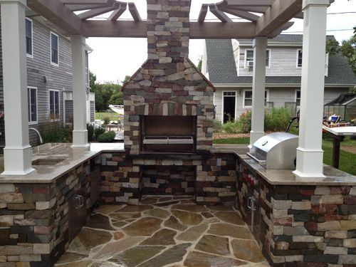 Outdoor kitchen with stone walls, fireplace, grill, and pergola in a backyard setting.