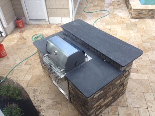 Outdoor kitchen with a grill, stone facade, and dark countertop. A hose and bucket are visible.