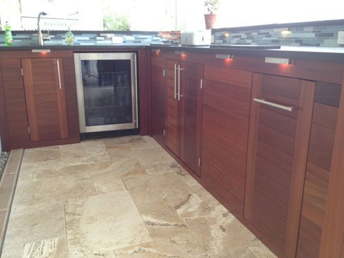 Outdoor kitchen with brown wooden cabinets, a stainless steel refrigerator, and stone tile flooring.