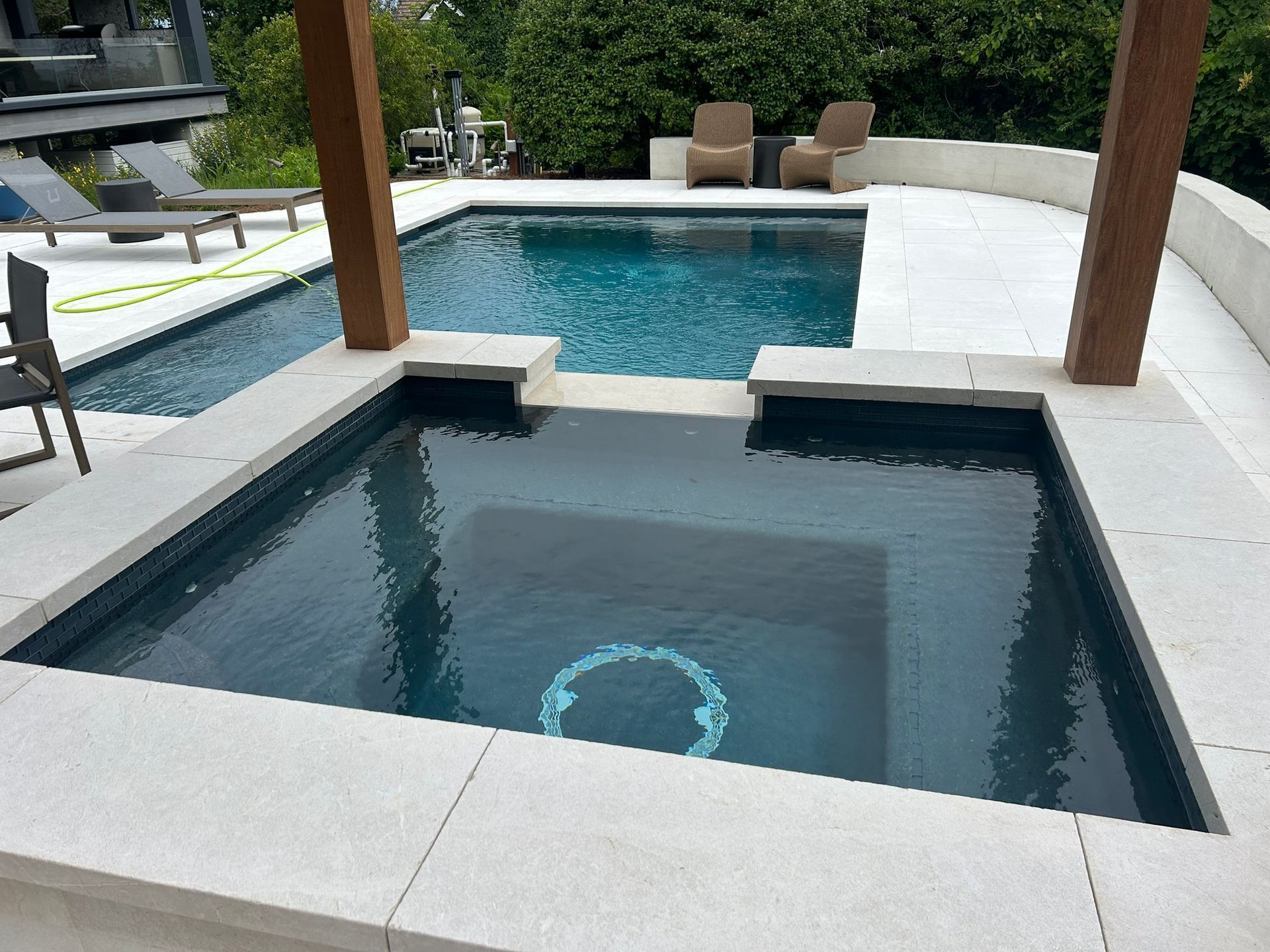 Pool and spa with wood pergola, surrounded by white stone. Water is dark blue.