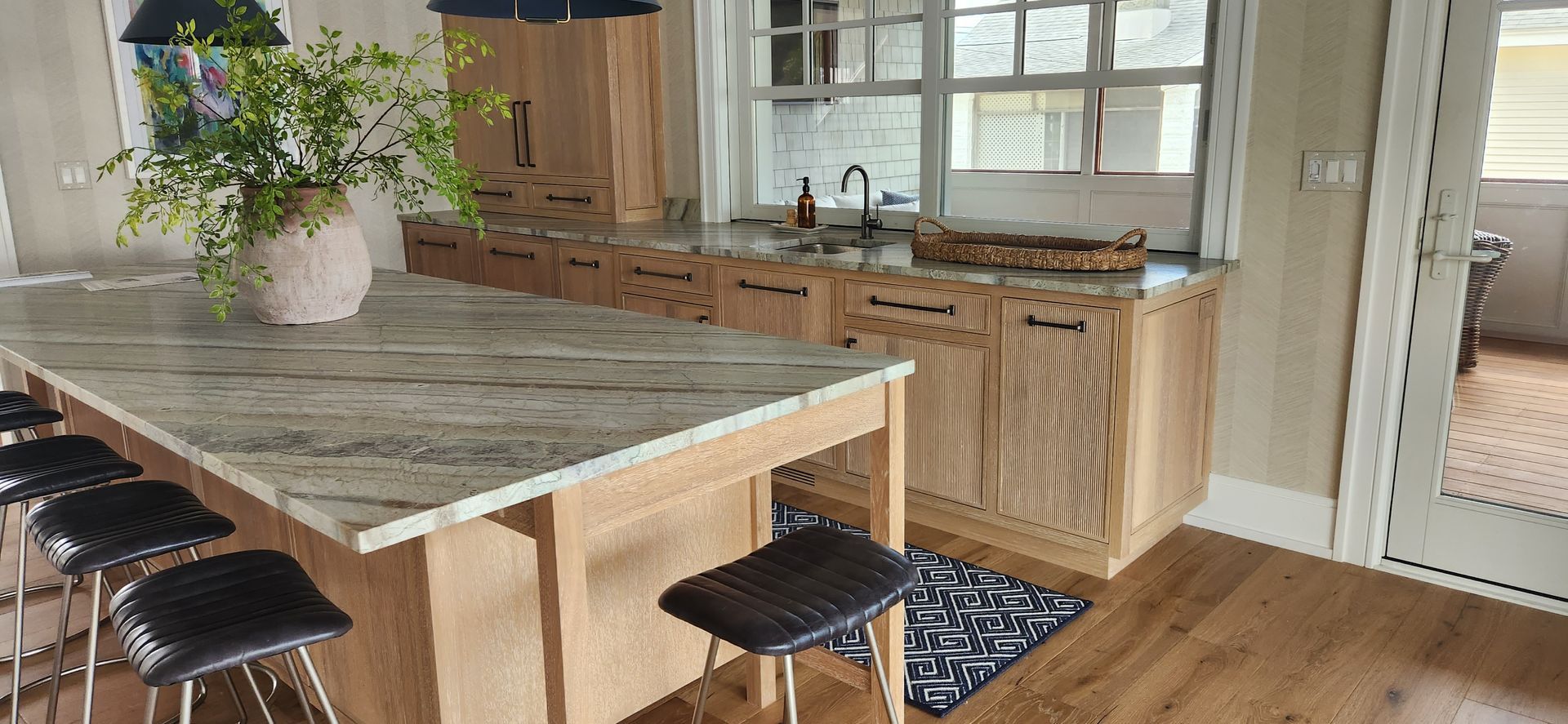Kitchen with light wood cabinets, granite countertops, and island with stools. Large windows in the background.