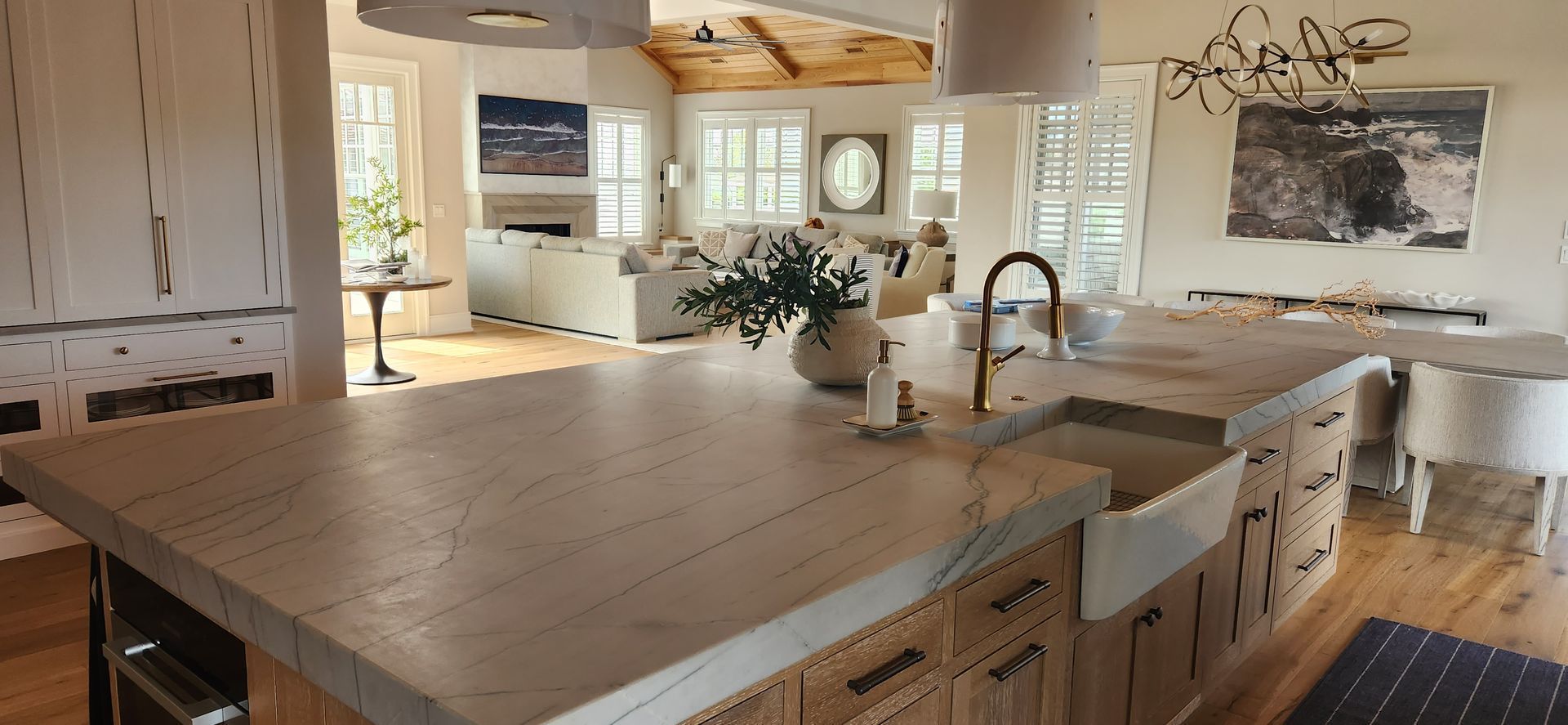 A light-filled kitchen with a large island and farmhouse sink. A living area with a sofa, artwork, and windows is in the background.