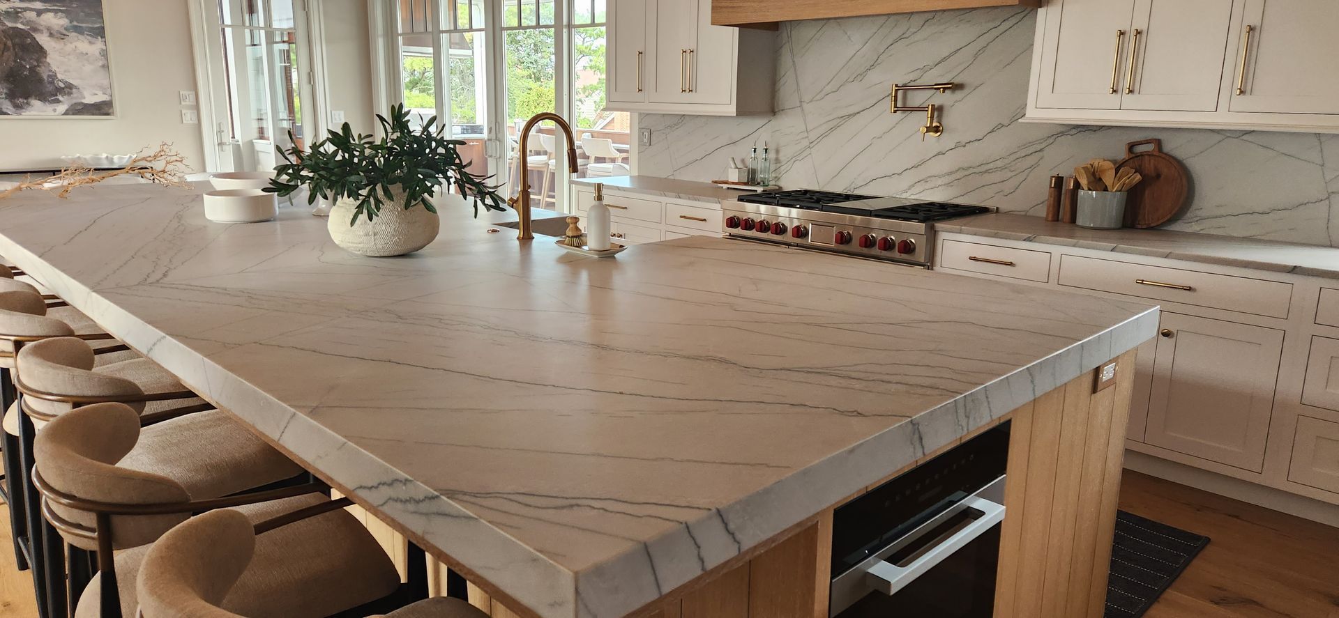 Kitchen island with white countertops, seating, and gold faucet, with greenery and light cabinets.
