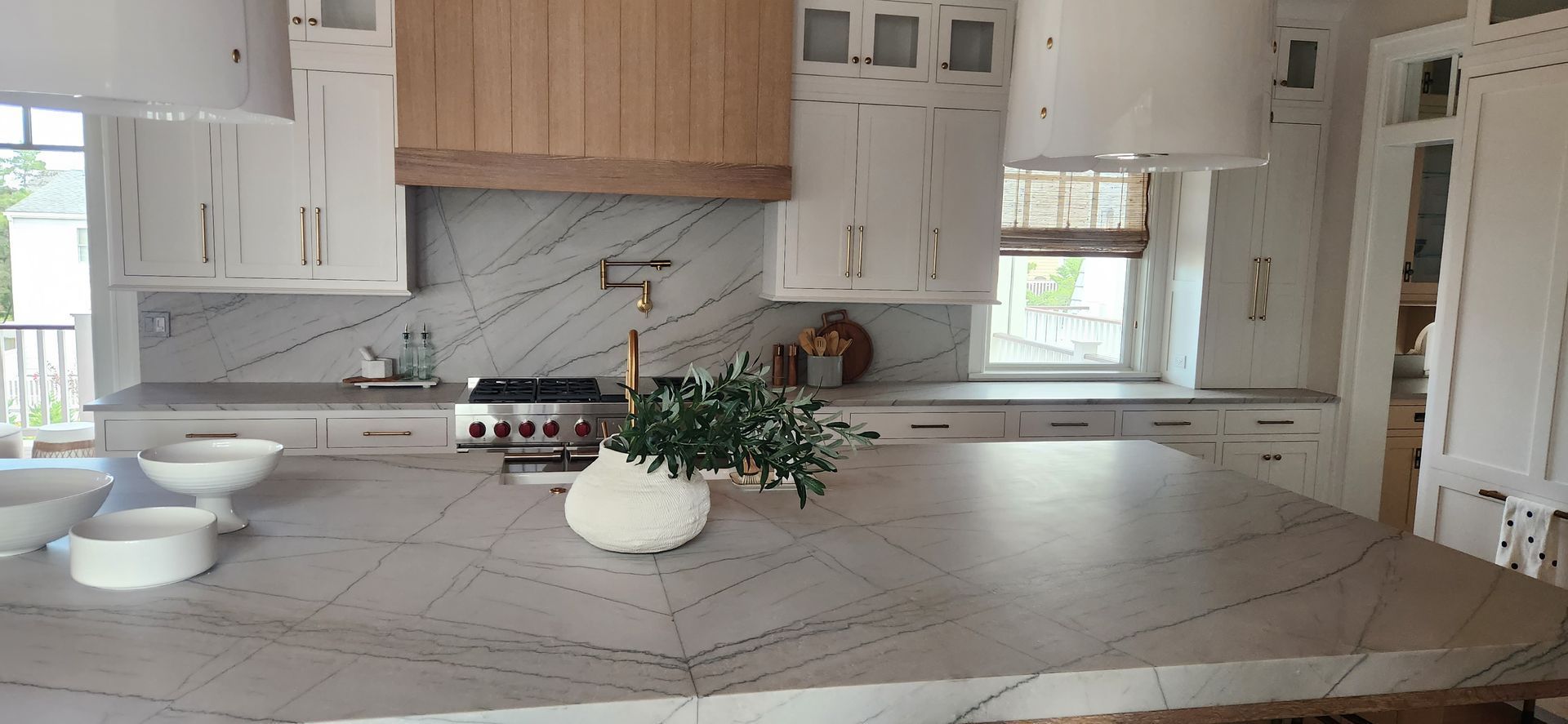 A kitchen with a large island, white cabinets, marble backsplash, and a wooden hood.