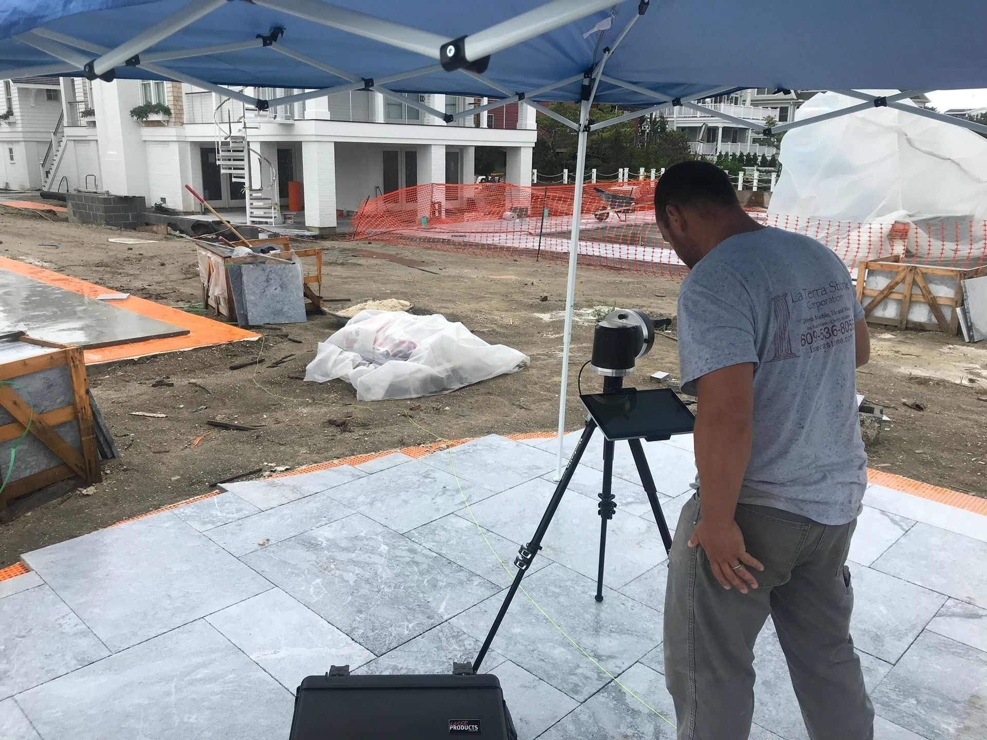 Man using a tripod-mounted camera under a canopy at a construction site.