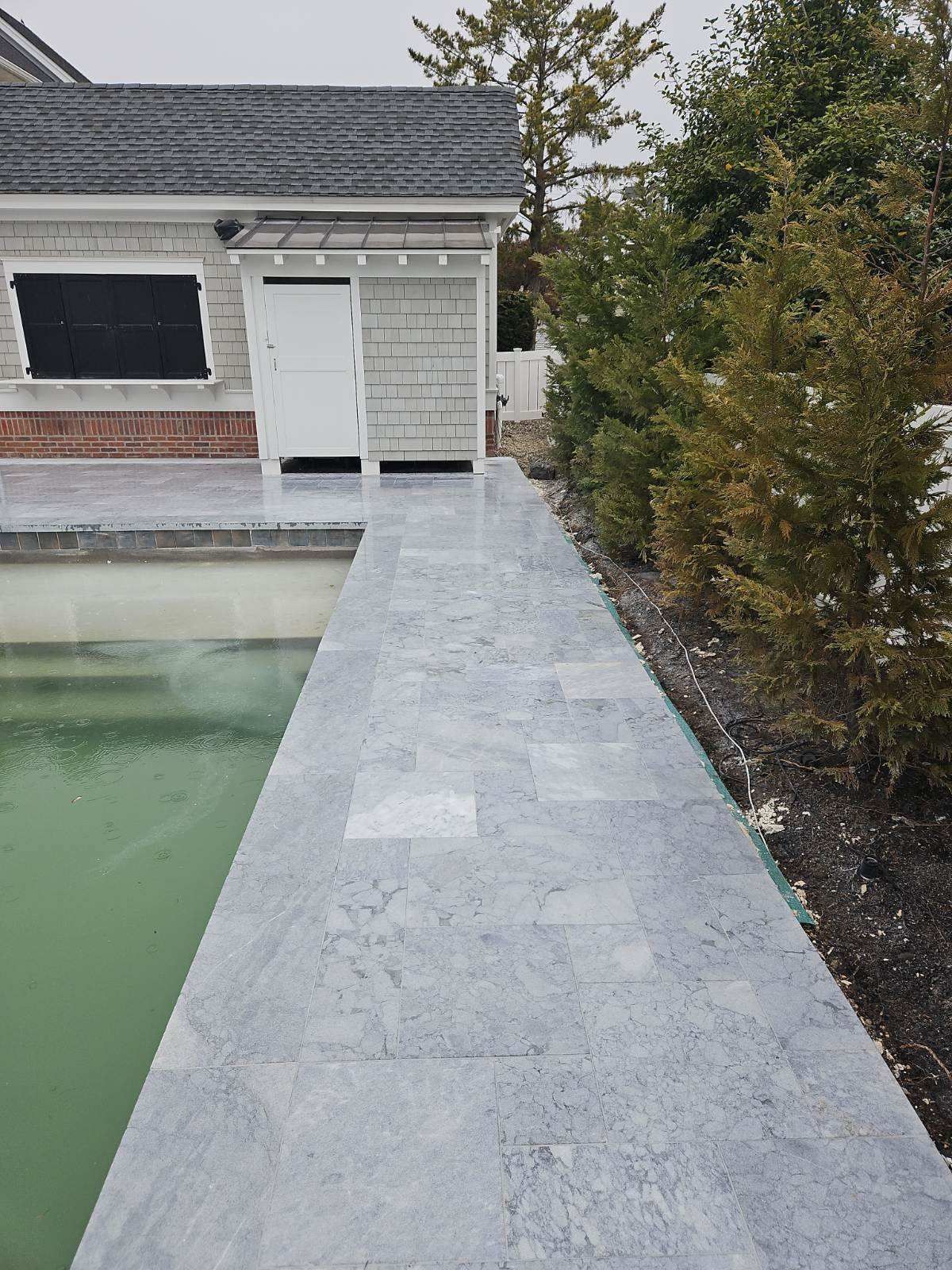 Stone walkway alongside a pool, leading to a light gray building with a white door.