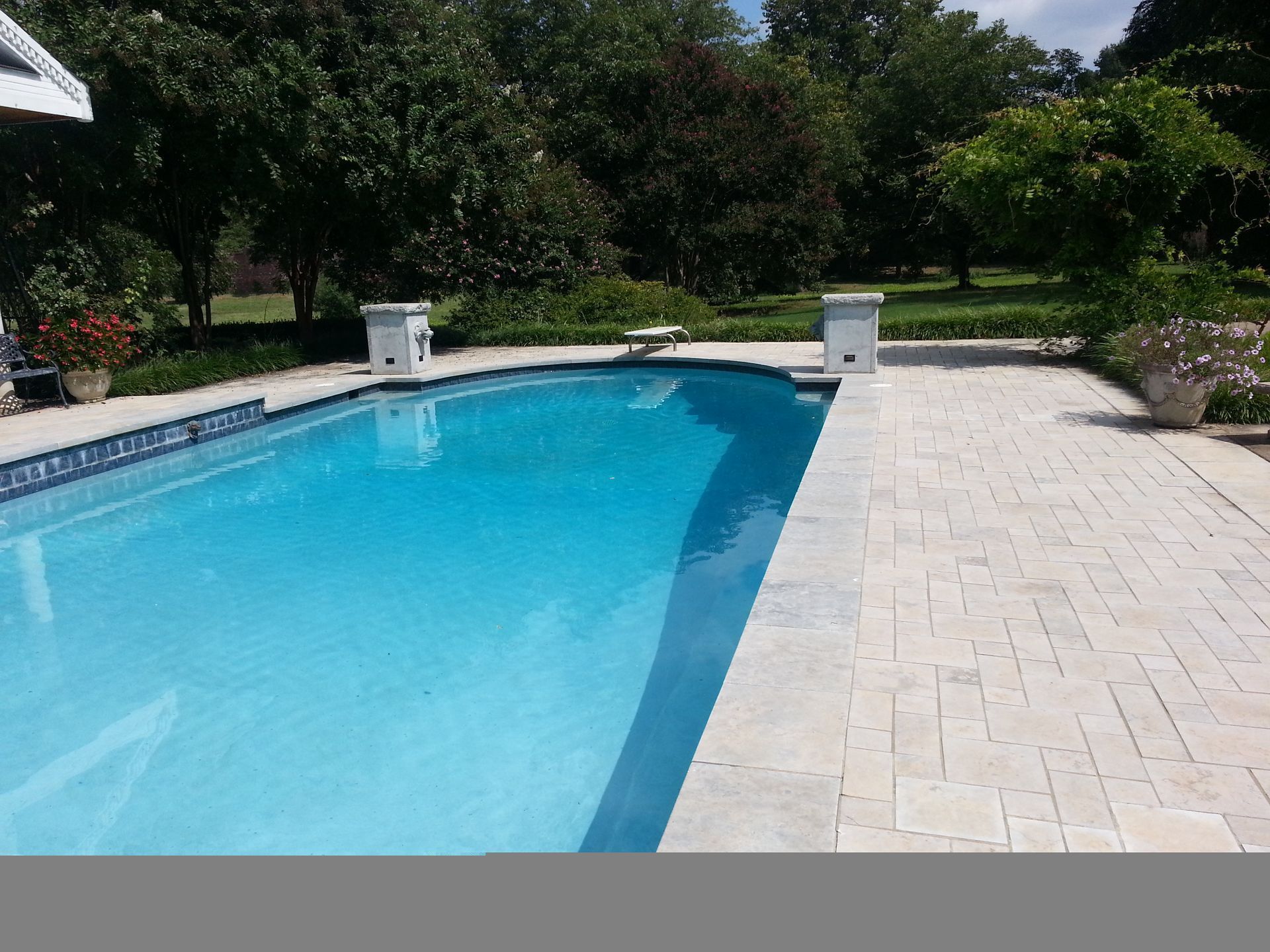 Outdoor swimming pool with blue water and light stone patio surrounded by trees.