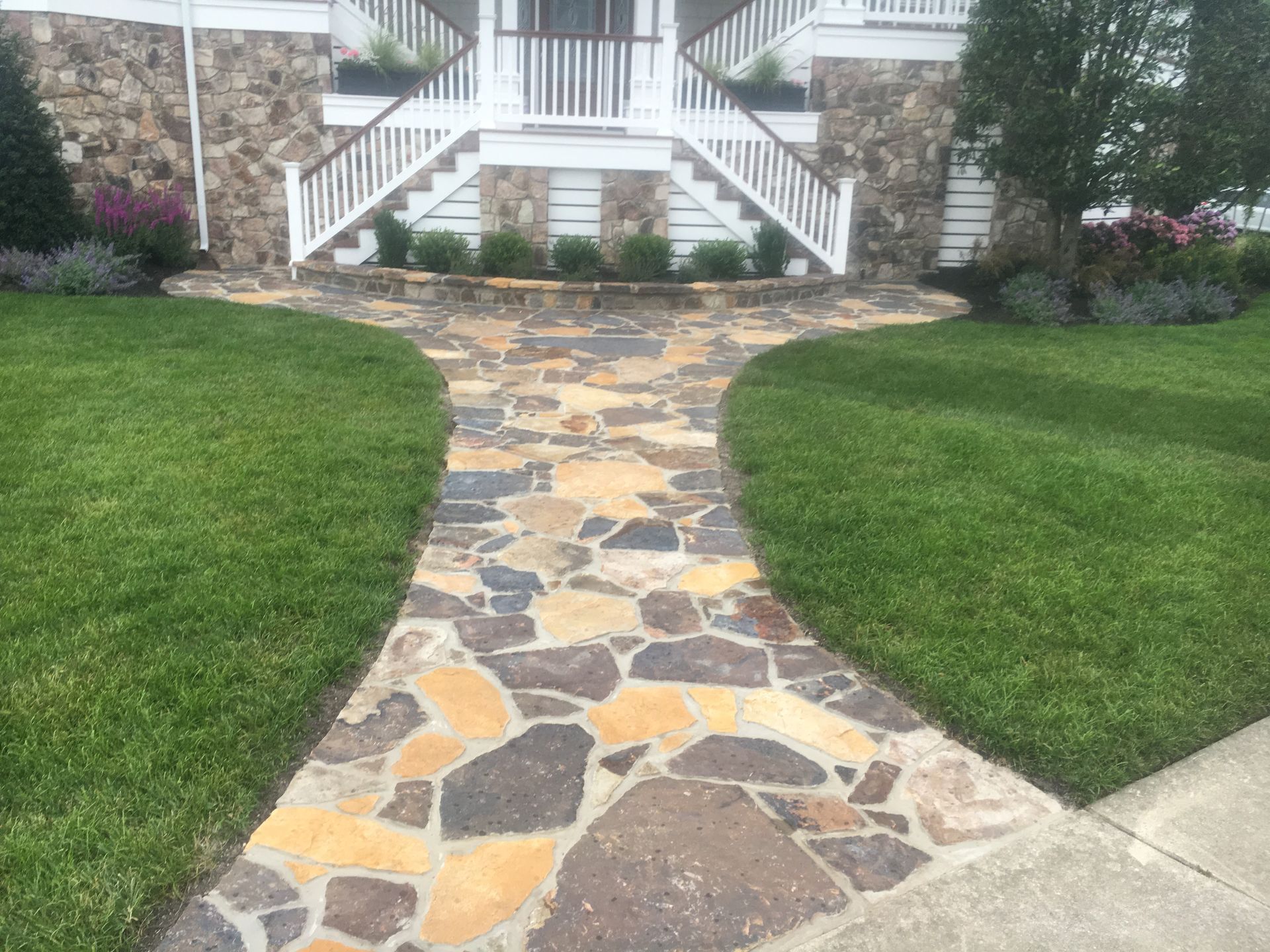 Stone pathway leading to a house with a white staircase, flanked by green grass and landscaping.
