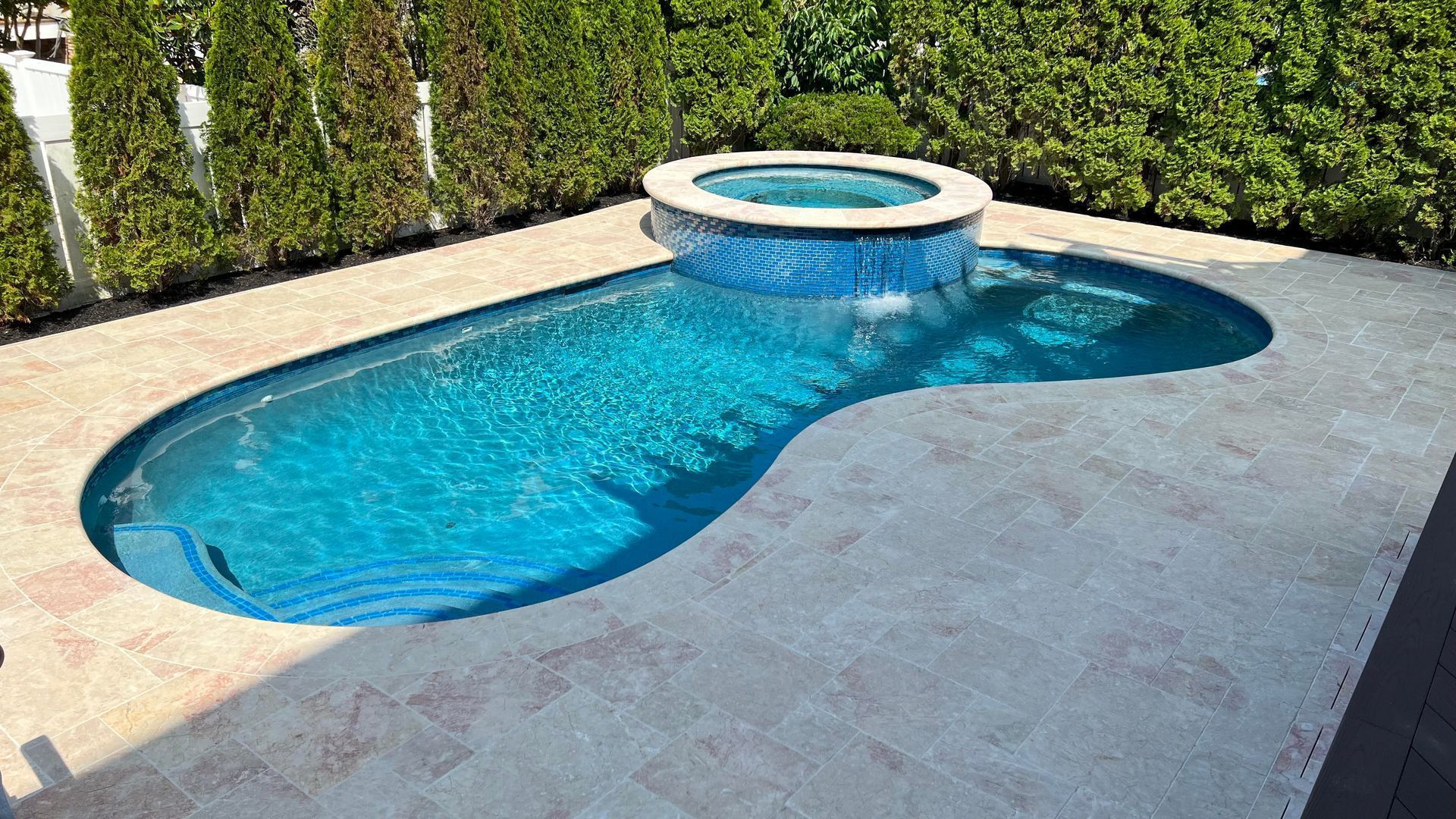 Swimming pool with attached raised hot tub, turquoise water, surrounded by beige stone, lush green bushes in the background.