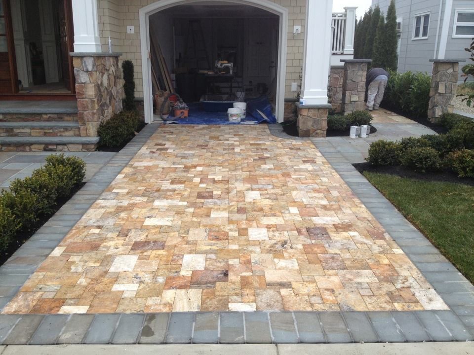 Driveway paved with tan stone tiles, bordered by gray pavers and greenery, leading to a garage entrance.