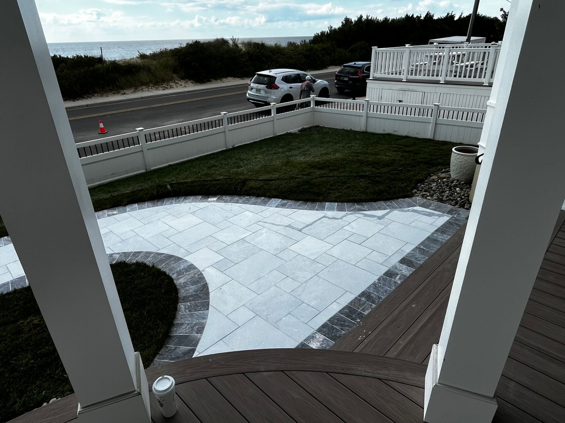 View from a porch overlooking a paved patio, grassy yard, road, and ocean.