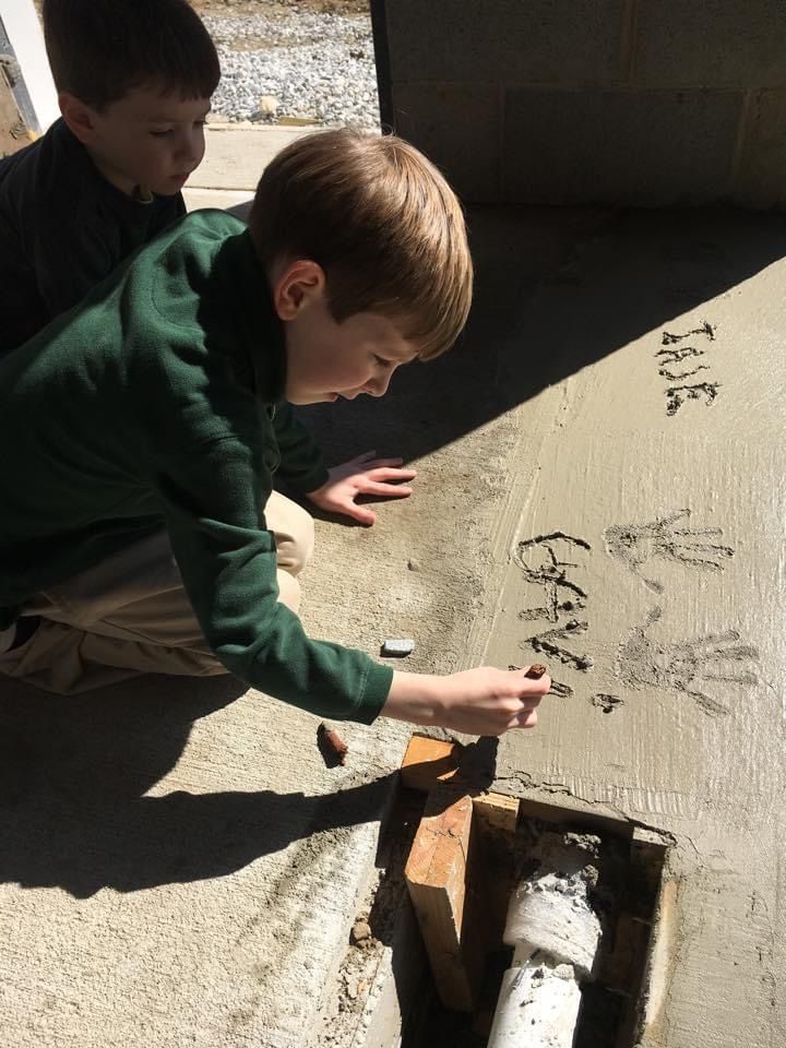 Two boys writing in wet concrete outside. One kneels, writing; the other looks on. The sun casts a shadow.